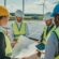 A group of project managers discussing plans at a construction site with wind turbines and solar panels in the background.