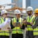 A group of business leaders and engineers discussing plans at a large construction site with cranes and bridges in the background.