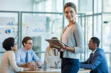 A young woman holding a tablet talks with colleagues around a conference table in a modern office.